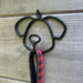 A black metal dog-shaped wall hook with a red and black plaid leash hanging from it, set against a rustic wooden plank background.