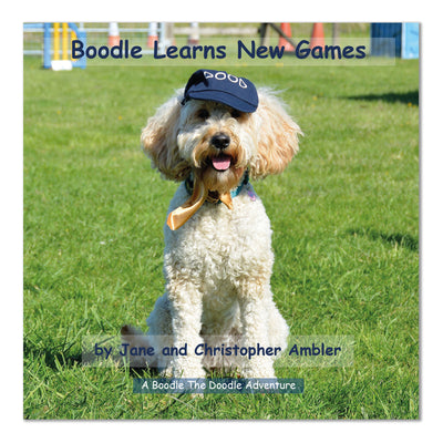 Front cover of the book Boodle Learns New Games which features a photograph of a smiley Golden Doodle with its tongue out, wearing a blue baseball cap sat on grass with agility games in the background. The cover also has the authors names Jane and Christopher Ambler and the words A Boodle The Doodle Adventure on it.