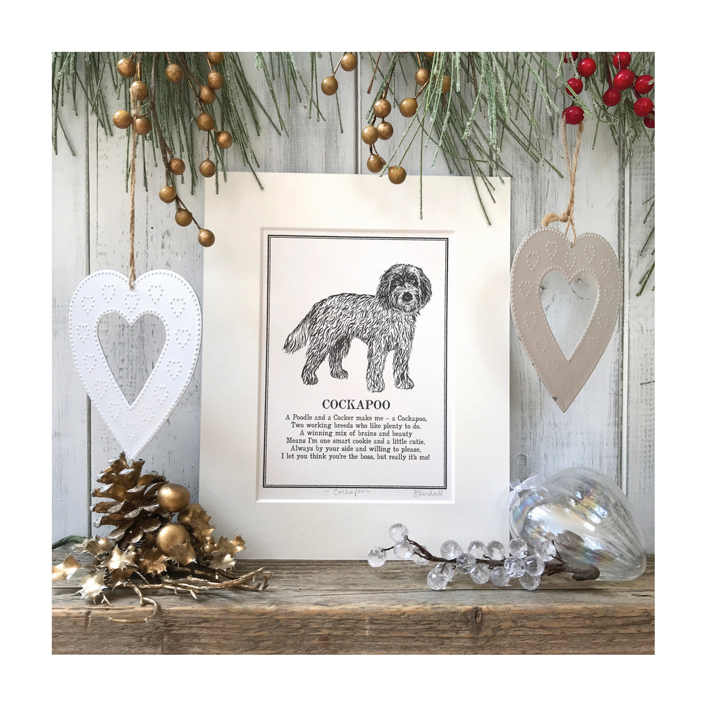 A print featuring a black and white line illustration of a Cockapoo with a poem about Cockapoos underneath. The print is in a cream mount and sat on a wooden surface with festive decorations around it.