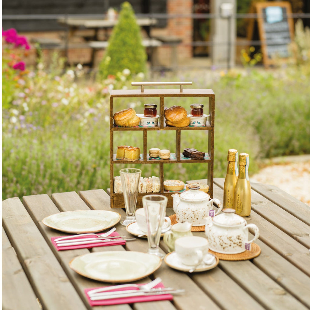 A photograph of the afternoon tea in a miniature wooden tiered box with a silver metal handle. The display box holds scones and sandwiches and other miniature assorted afternoon tea snacks with teapots, teacups, glasses, prosecco and plates in front. All laid out on a table outdoors with colourful flowers and the Grange restaurant in the background.