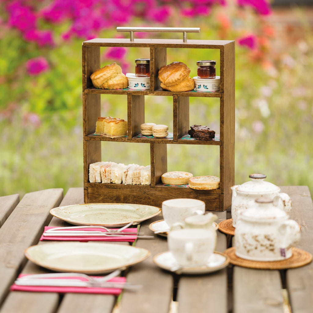 A photograph of the afternoon tea in a miniature wooden tiered box with a silver metal handle. The display box holds scones and sandwiches and other miniature assorted afternoon tea snacks with teapots, teacups and plates in front. All laid out on a table at The Grange outdoors with colourful flowers in the background.