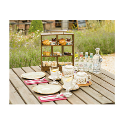 A photograph of the afternoon tea in a miniature wooden tiered box with a silver metal handle. The display box holds scones and sandwiches and other miniature assorted afternoon tea snacks with teapots, teacups, glasses and an example of the gin and tonic and plates in front. All laid out on a table outdoors with colourful flowers and the Grange restaurant in the background.