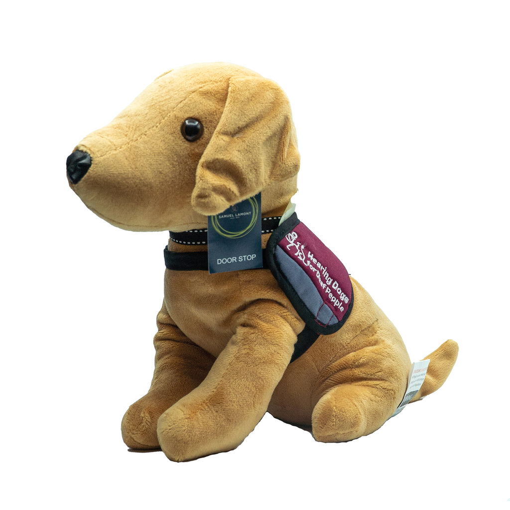 Side view of a doorstop made to look like a cuddly yellow Labrador wearing a Hearing Dogs burgundy jacket. The dog has brown eyes, a black nose and is wearing a dog collar with a tag that says 'Door Stop' on it.
