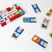 Dog Domino game set with a red and white box and multiple rectangular domino tiles featuring different dog breeds on colorful blue, orange, yellow, and light blue backgrounds, arranged on a white surface.