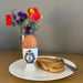 Egg in an egg cup with a dog design, next to toast and a fork on a plate, with flowers in the background.