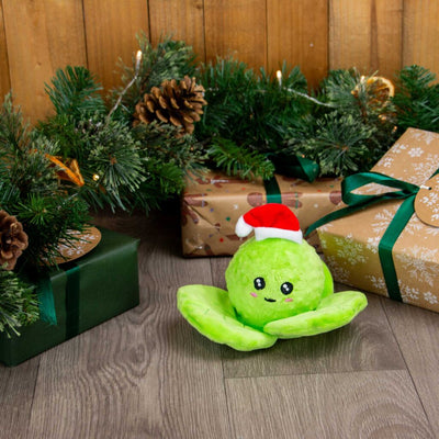 A bright green plush brussel sprout toy wearing a small red Santa hat, posed on a wood floor with Christmas greenery, pinecones, and wrapped gift boxes in the background.