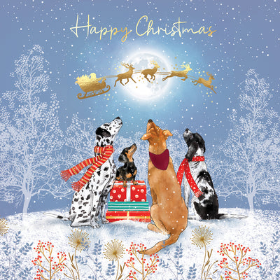 A festive Christmas illustration featuring four dogs sitting in the snow beside wrapped gifts, looking up at Santa Claus flying in a sleigh under a full moon. The scene is decorated with snow-covered trees, gold stars, and the words 'Happy Christmas' at the top.