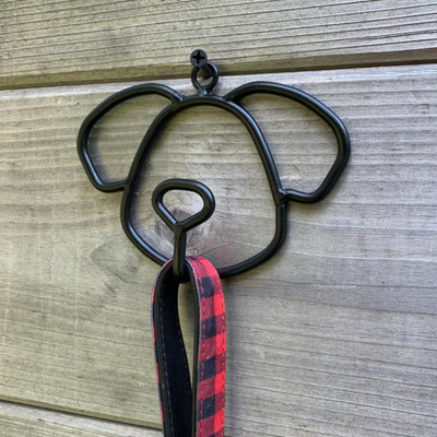 A black metal dog-shaped wall hook with a red and black plaid leash hanging from it, set against a rustic wooden plank background.
