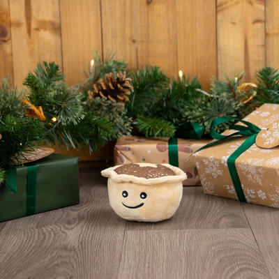 Plush mince pie toy on a wooden floor with Christmas decorations and presents.