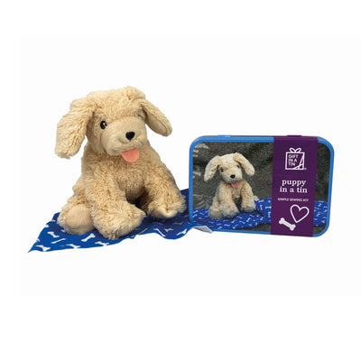 A cute small soft dog toy, with its tongue stuck out, sat on a blue blanket with bone shapes on it next to a tin in which it came. The metal tin has a picture of the same soft dog toy on it.