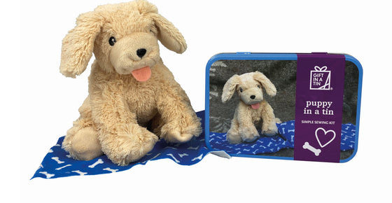 A cute small soft dog toy, with its tongue stuck out, sat on a blue blanket with bone shapes on it next to a tin in which it came. The metal tin has a picture of the same soft dog toy on it.