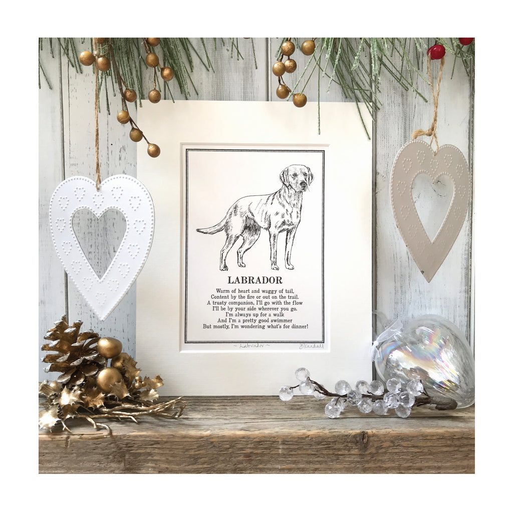 A print featuring a black and white line illustration of a Labrador with a poem about Labradors underneath. The print is in a cream mount and sat on a wooden surface with festive decorations around it.