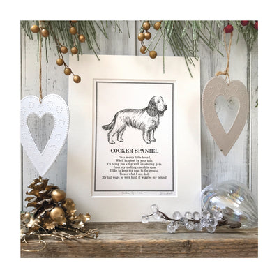 A print featuring a black and white line illustration of a Cocker Spaniel with a poem about Spaniels underneath. The print is in a cream mount and sat on a wooden surface with festive decorations around it.