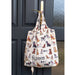 Shopper bag hanging on a gold metal coloured door handled on a dark grey door. The shopper bag features sketch illustrations of dogs of different sizes, colours and breeds all wearing the official Hearing Dogs jacket. The bag appears to be full.