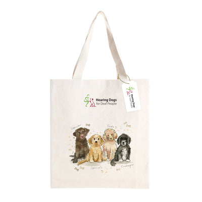 A front on view of the tote bag with the handles pointing upwards. The beige tote bag has illustrations of 4 dogs on the centre of the bag, from left to right, a Labrador, Spaniel, Poodle and Cockapoo. The Hearing Dogs logo is positioned on the centre top of the bag, with a Hearing Dogs label attached to the handles.