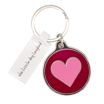 Close up photo of Dog Tag or Keyring. Attached to the metal loop is a Red coloured circular keyring with a Pink Heart design printed on it. Also attached to the metal loop is a silver rectangular dog tag which reads: 'the little dog laughed'. 