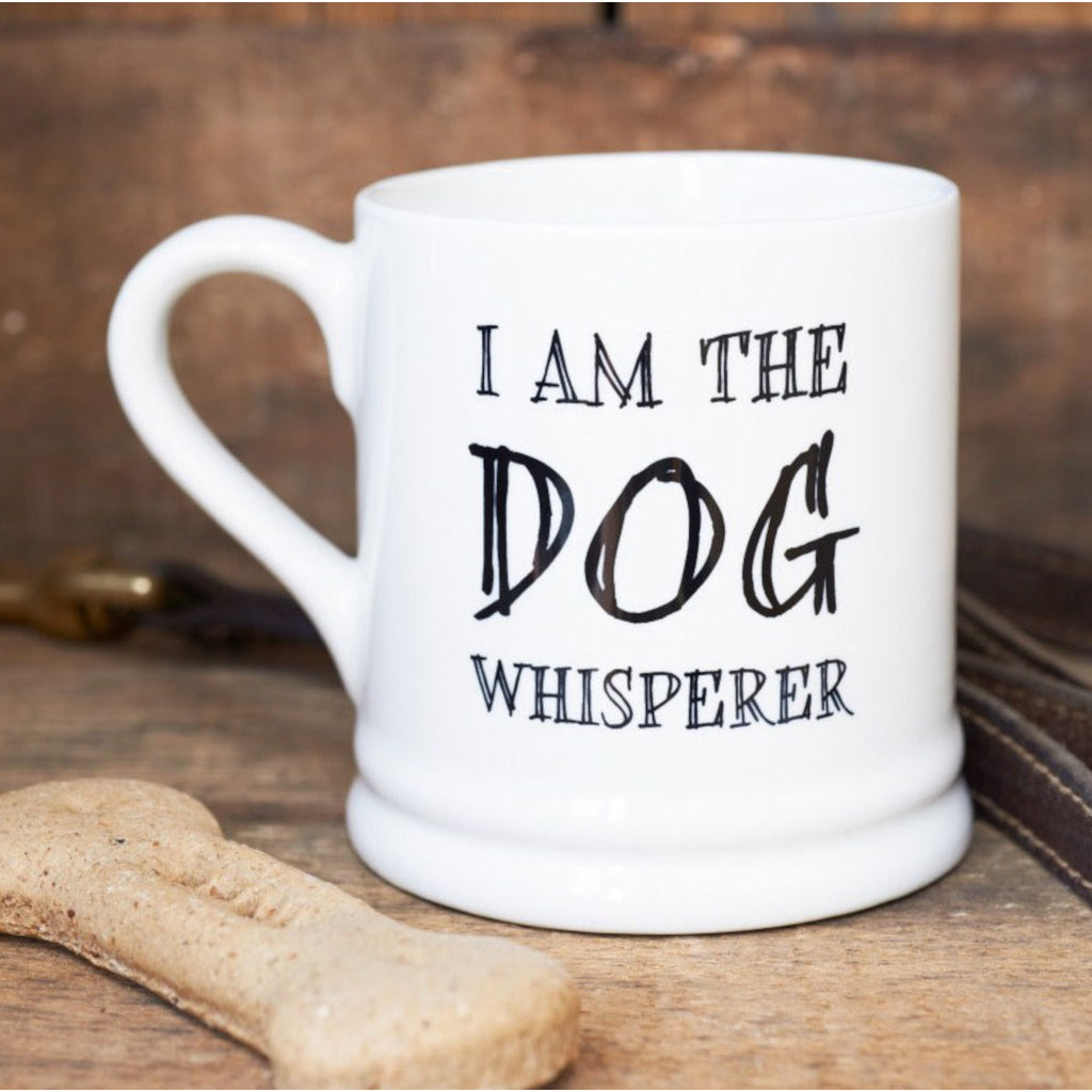 Image shows a white / cream mug, with black font on the front of mug saying 'I am the dog whisperer'. The mug is sat on a table top, next to a small brown bone.