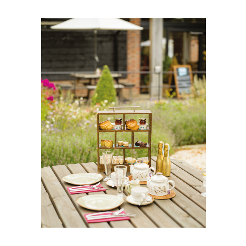 A photograph of the afternoon tea in a miniature wooden tiered box with a silver metal handle. The display box holds scones and sandwiches and other miniature assorted afternoon tea snacks with teapots, teacups, glasses, prosecco and plates in front. All laid out on a table outdoors with colourful flowers and the Grange restaurant in the background.