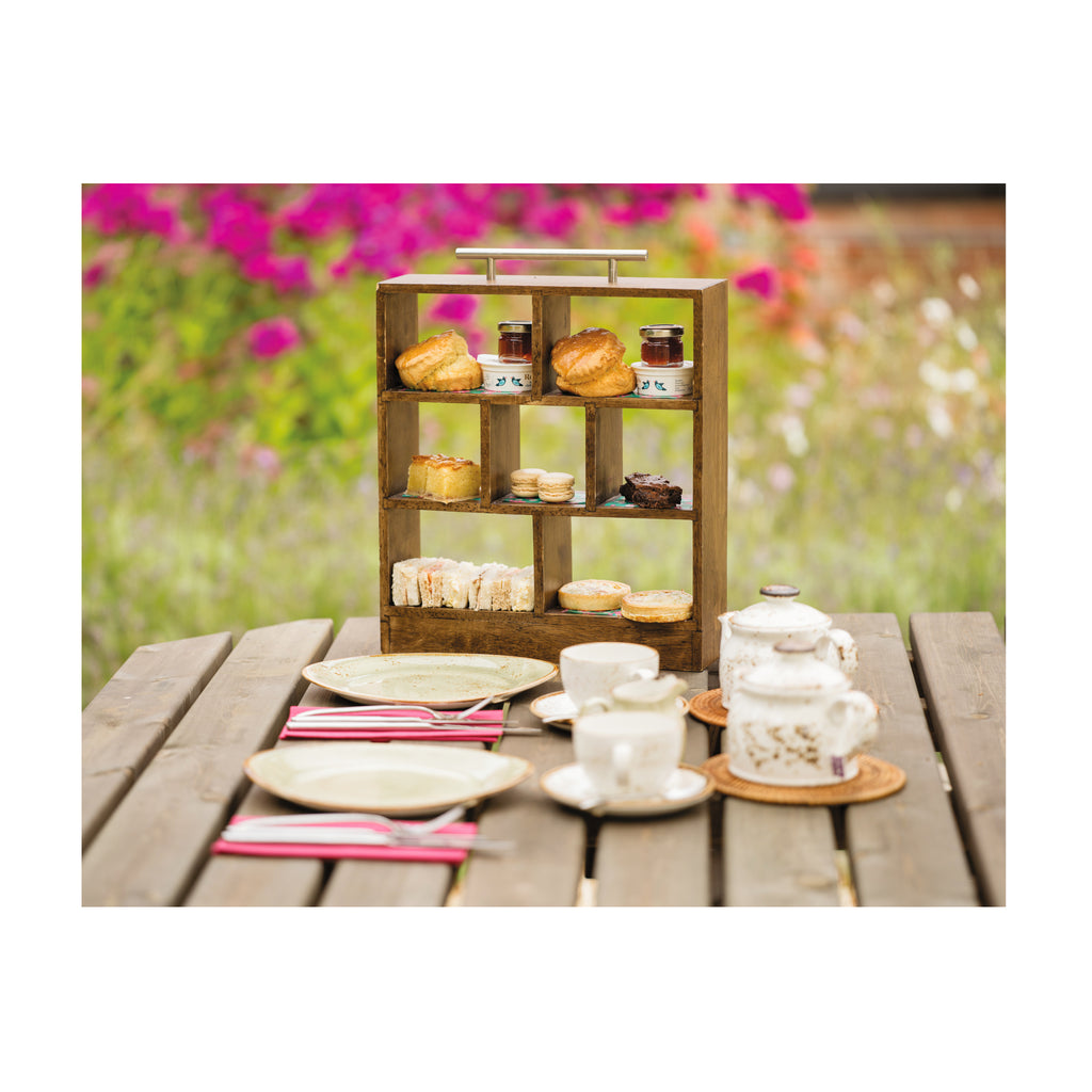 A photograph of the afternoon tea in a miniature wooden tiered box with a silver metal handle. The display box holds scones and sandwiches and other miniature assorted afternoon tea snacks with teapots, teacups and plates in front. All laid out on a table at The Grange outdoors with colourful flowers in the background.
