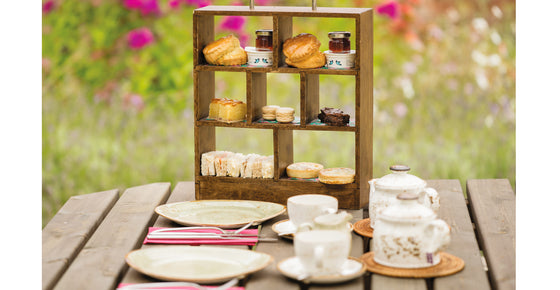 A photograph of the afternoon tea in a miniature wooden tiered box with a silver metal handle. The display box holds scones and sandwiches and other miniature assorted afternoon tea snacks with teapots, teacups and plates in front. All laid out on a table at The Grange outdoors with colourful flowers in the background.