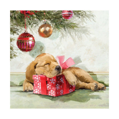 The front cover of a festive greeting card. The image on the front of the card is an illustrated golden Labrador puppy asleep under the Christmas tree with its head resting on a red Christmas present with a red bow. There are red and gold baubles hanging over the puppy from the tree.